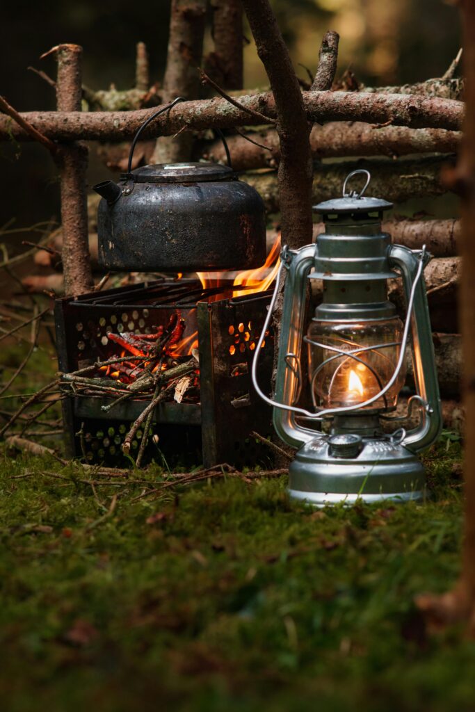 Rustic outdoor campfire setting with a kettle and a glowing lantern on mossy ground.